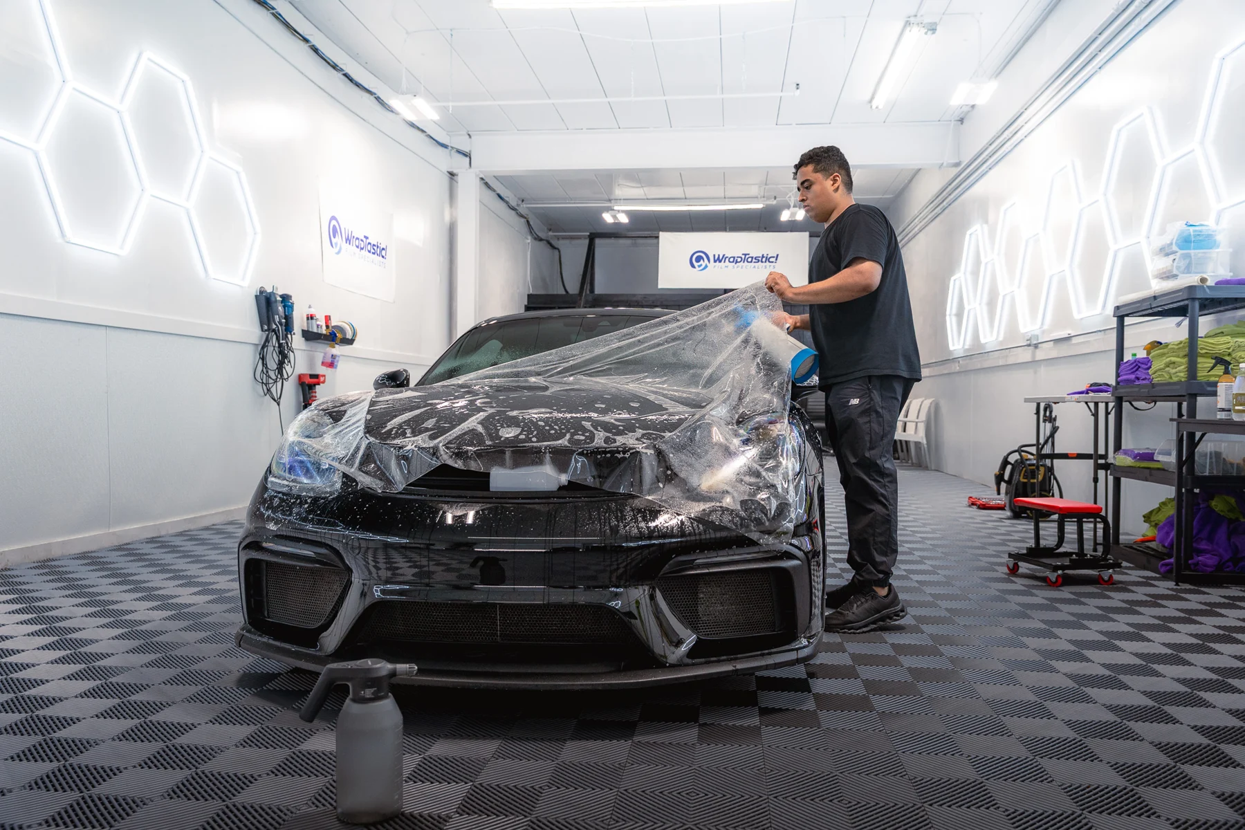 WrapTastic technician fitting clear paint-protection film to a black Porsche in the studio